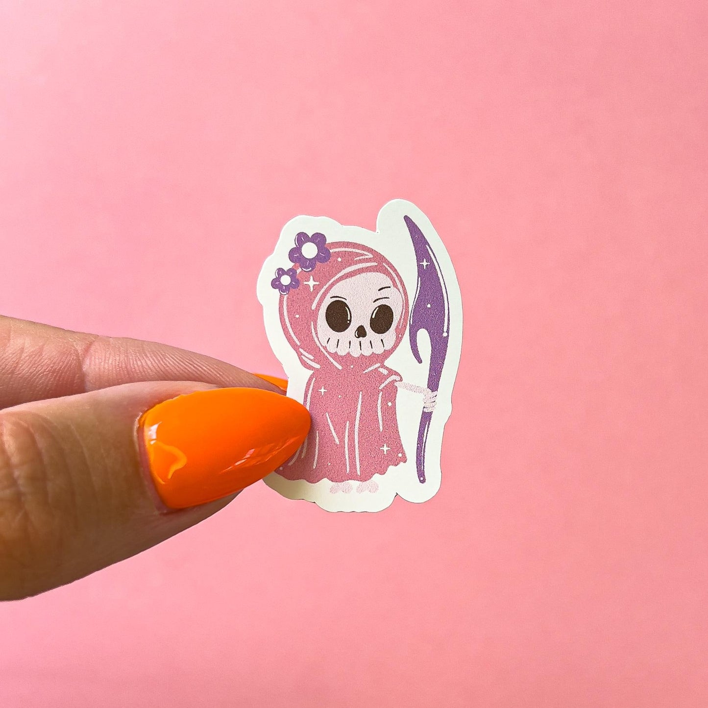 A vinyl decal sticker of a girly grim reaper character. The grim reaper has a white skeleton face. She is wearing a pink cape/robe with purple flowers on the hood. She is holding a purple scythe.