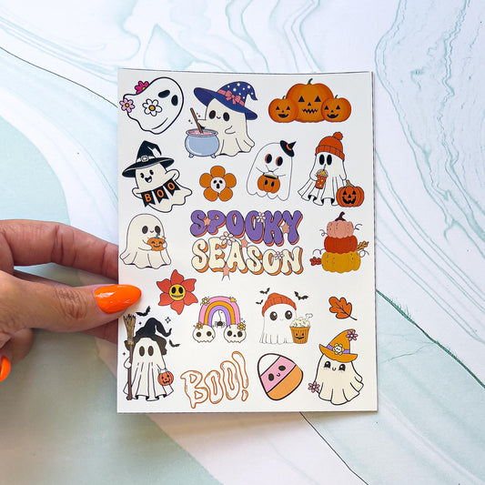 A sheet of Halloween themed stickers. The stickers include "SPOOKY SEASON" in purple, orange, and cream styled text with flowers, jack-o-lantern pumpkins, cartoon candy corn, leaves, "BOO" in orange spooky text, and cute cartoon ghosts in various outfits, poses, with different objects. Some ghosts have a witches hat, are stirring a cauldron, holding a trick or treat bucket, holding a pumpkin, or holding a sign that says "BOO"