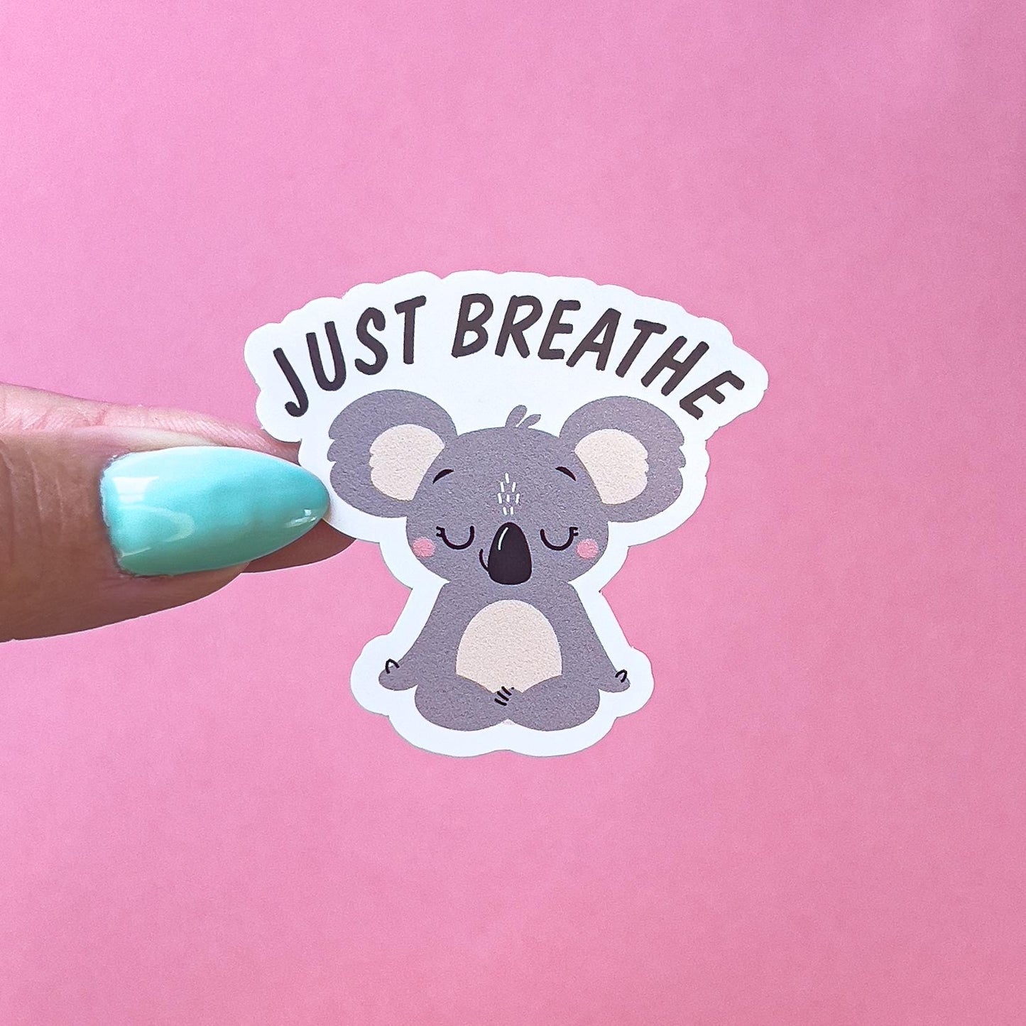A vinyl decal sticker of a grey koala  doing the Sukhasana, yoga pose. Above the koala black text reads “Just Breathe”. The koala has pink blushed cheeks.