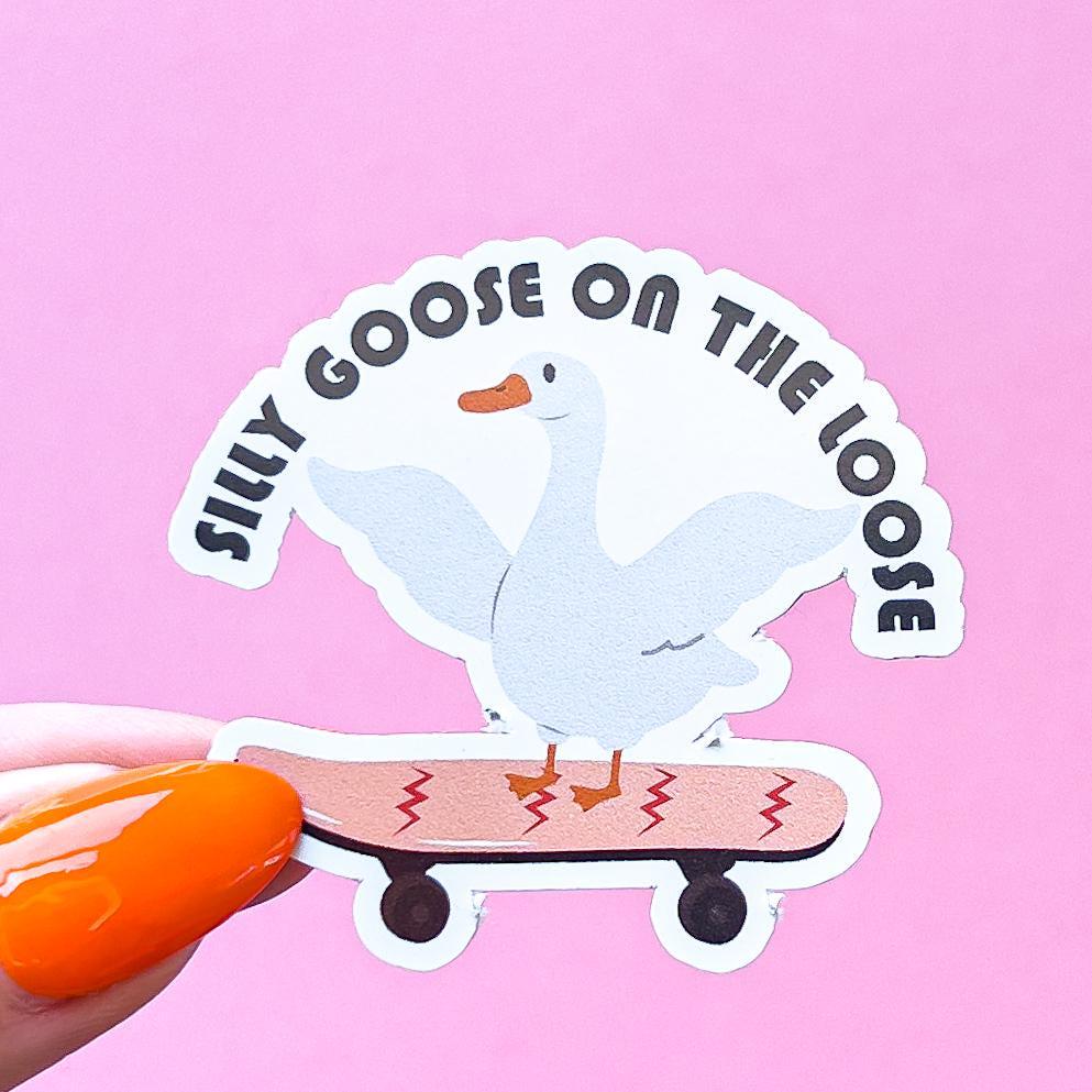 A vinyl decal sticker of a white goose with an orange beak is riding a skateboard. The skateboard is light red with red squiggly lines. The goose has its wings spread out. The text around the goose reads 'Silly Goose on the Loose'.