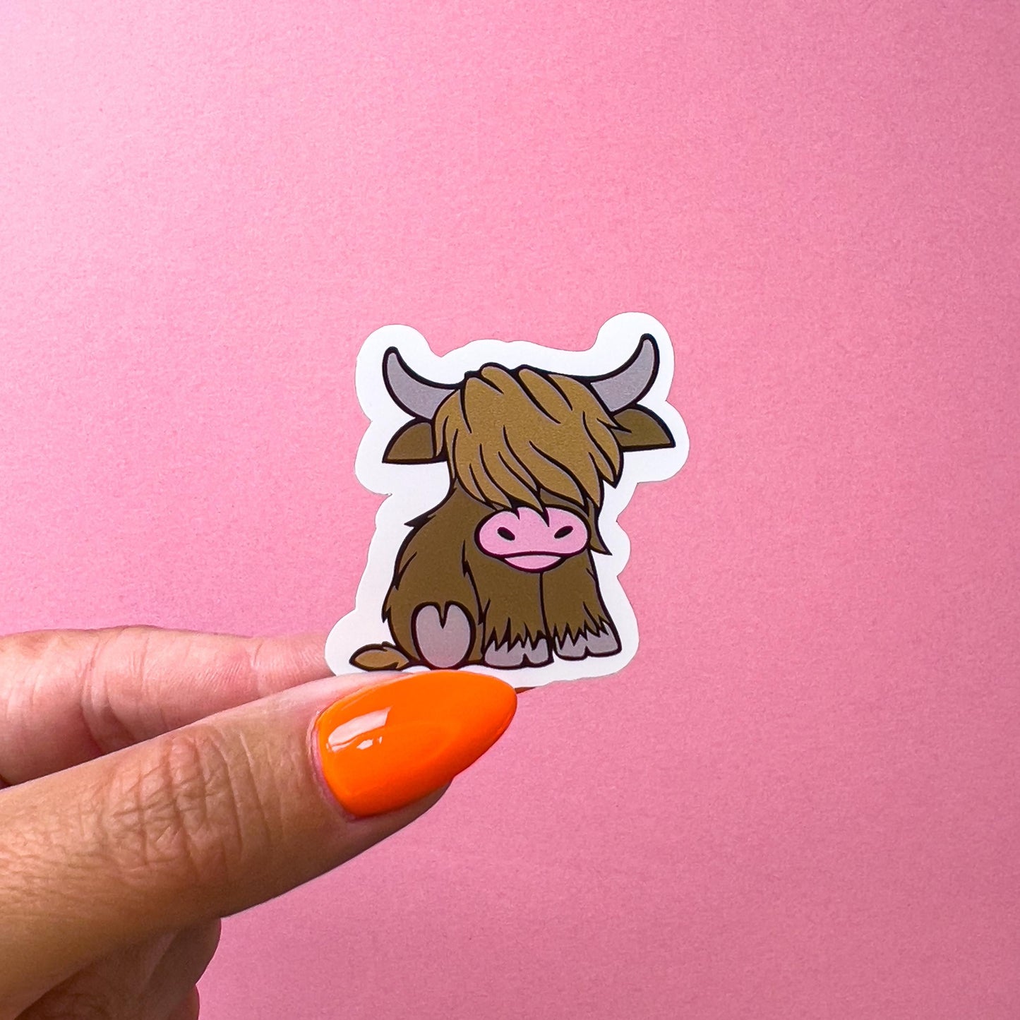 A vinyl decal sticker of a brown, cartoon highland cow with a pink nose. The cow is sitting down.