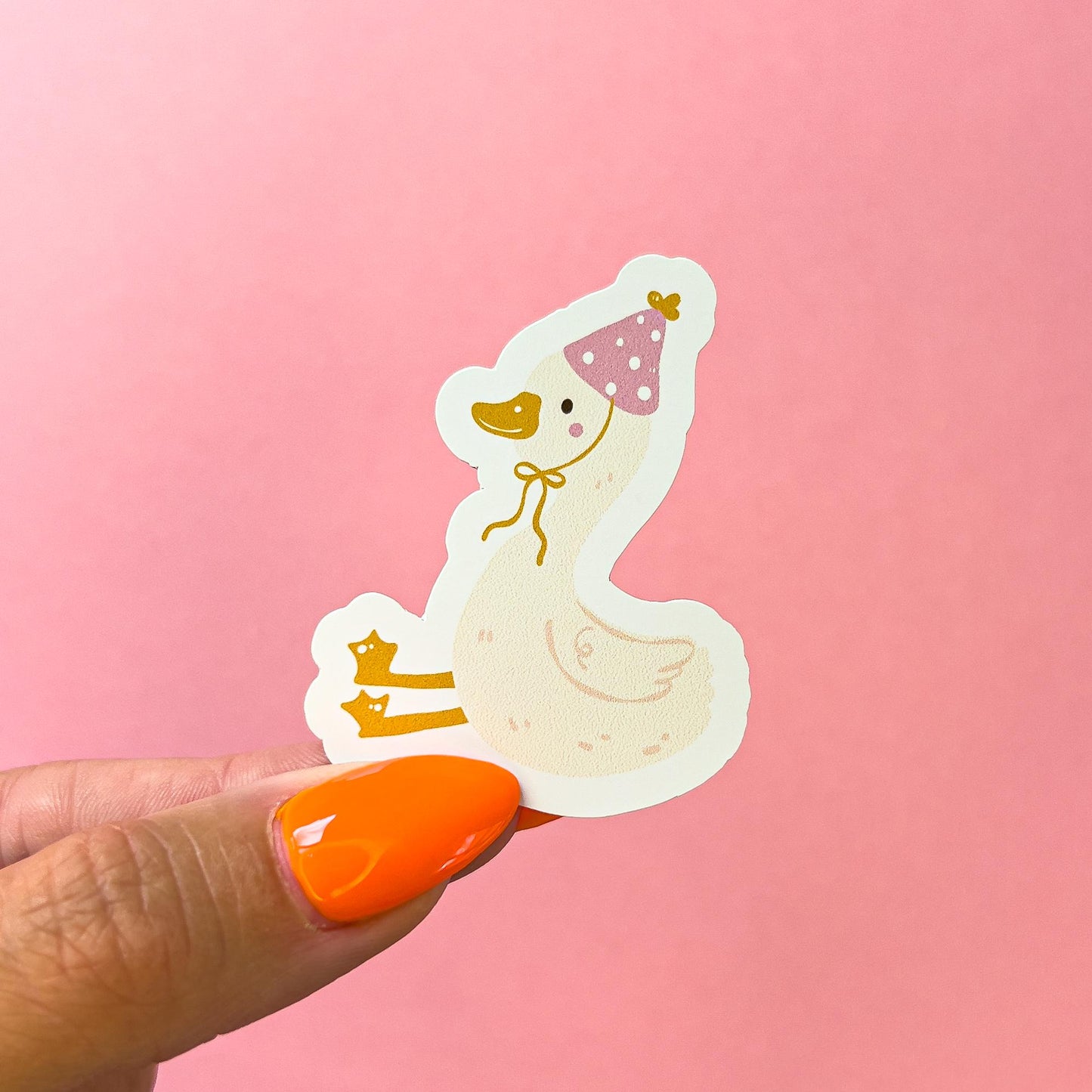 A sticker of a white goose wearing a pink party hat with white polka dots. The goose has a yellow beak and feet. The goose is sitting with its legs stretched out in front of him.