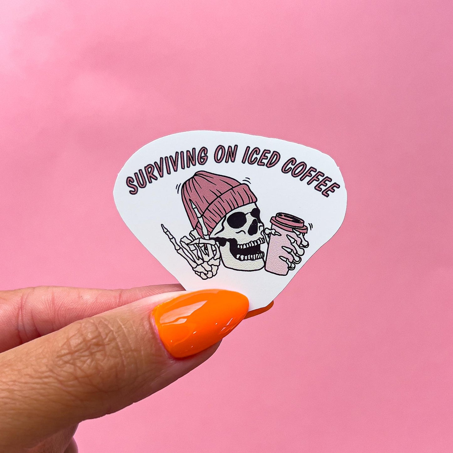 A vinyl decal sticker of a smiling skeleton wearing a mauve colored beanie, holding up the "rock on" hand symbol and holding a pink and mauve coffee cup. The text reads "Surviving on iced coffee".