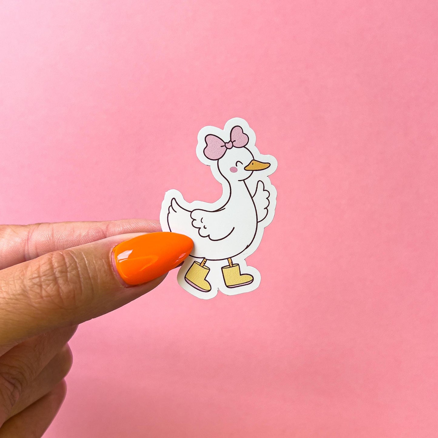 A sticker of a white duck/goose wearing yellow rain boots and a pink bow. The duck/goose is facing right with one wing up, waving. it also has a smiling face with blushing cheeks.