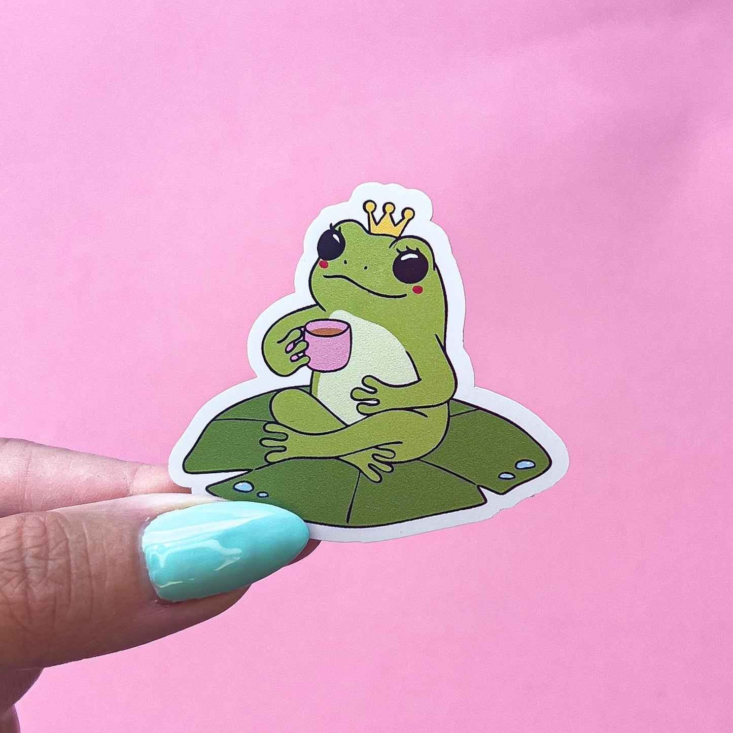 A vinyl decal sticker of a green, cartoon frog with blushed cheeks, wearing a crown. The frog is sitting criss cross on a lily pad and holding a pink coffee mug.