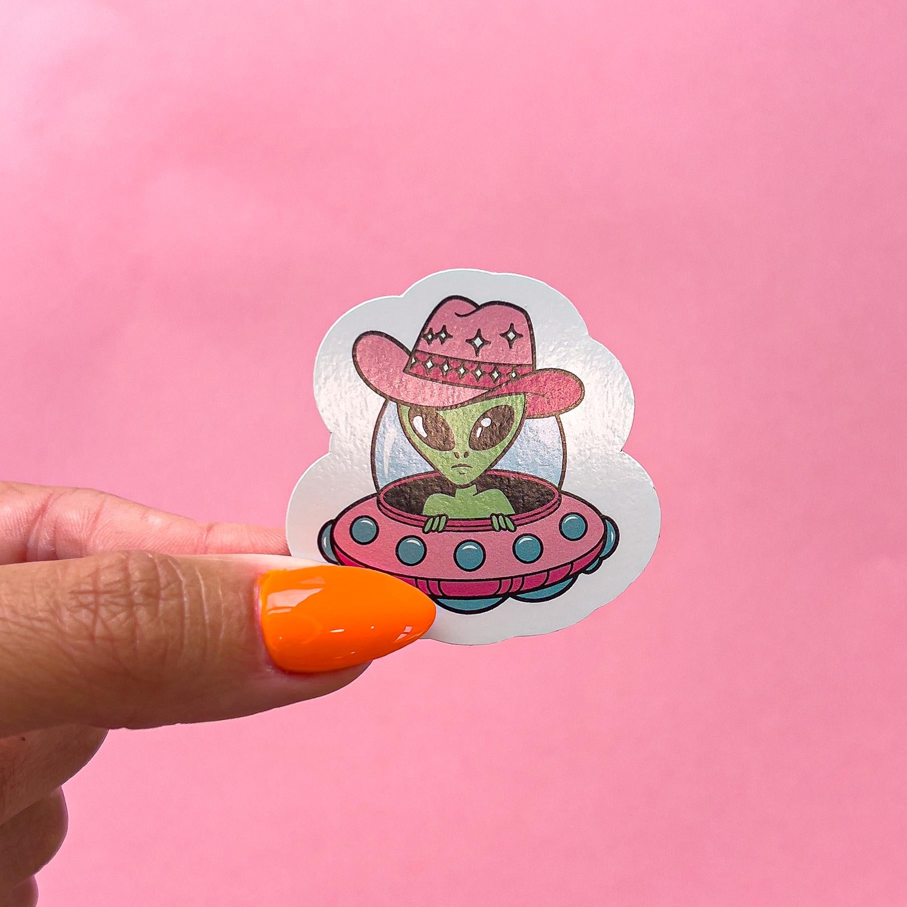 A vinyl decal sticker of a green alien wearing a pink cowboy hat and riding in a pink flying saucer spaceship.