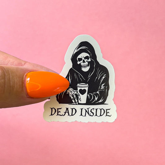 A black and white sticker of a hooded skeleton holding a coffee cup. the coffe cup has a black heart on it.  Black text reads  'DEAD INSIDE' is below the figure.