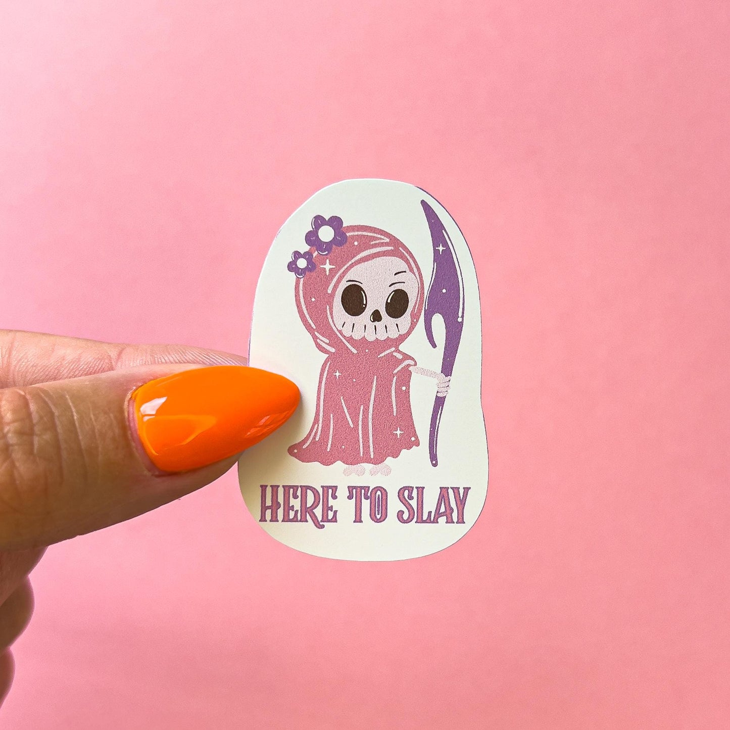 A vinyl decal sticker of a girly grim reaper character. The grim reaper has a white skeleton face. She is wearing a pink cape/robe with purple flowers on the hood. She is holding a purple scythe. reaper. The sticker says "HERE TO SLAY" underneath.