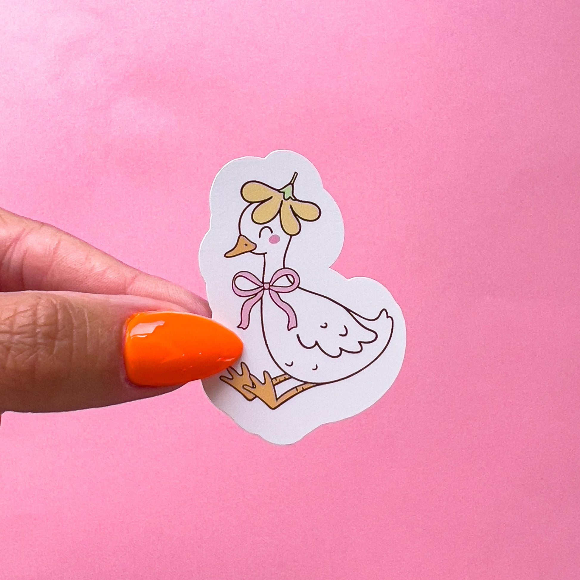 A vinyl decal sticker of a white goose/duck with yellow feet and beak. The duck/goose is sitting with its legs stretched out in front. The goose/duck is wearing a yellow flower as a hat and has a pink bow around its neck. The goose/duck is smiling with pink blushed cheeks.