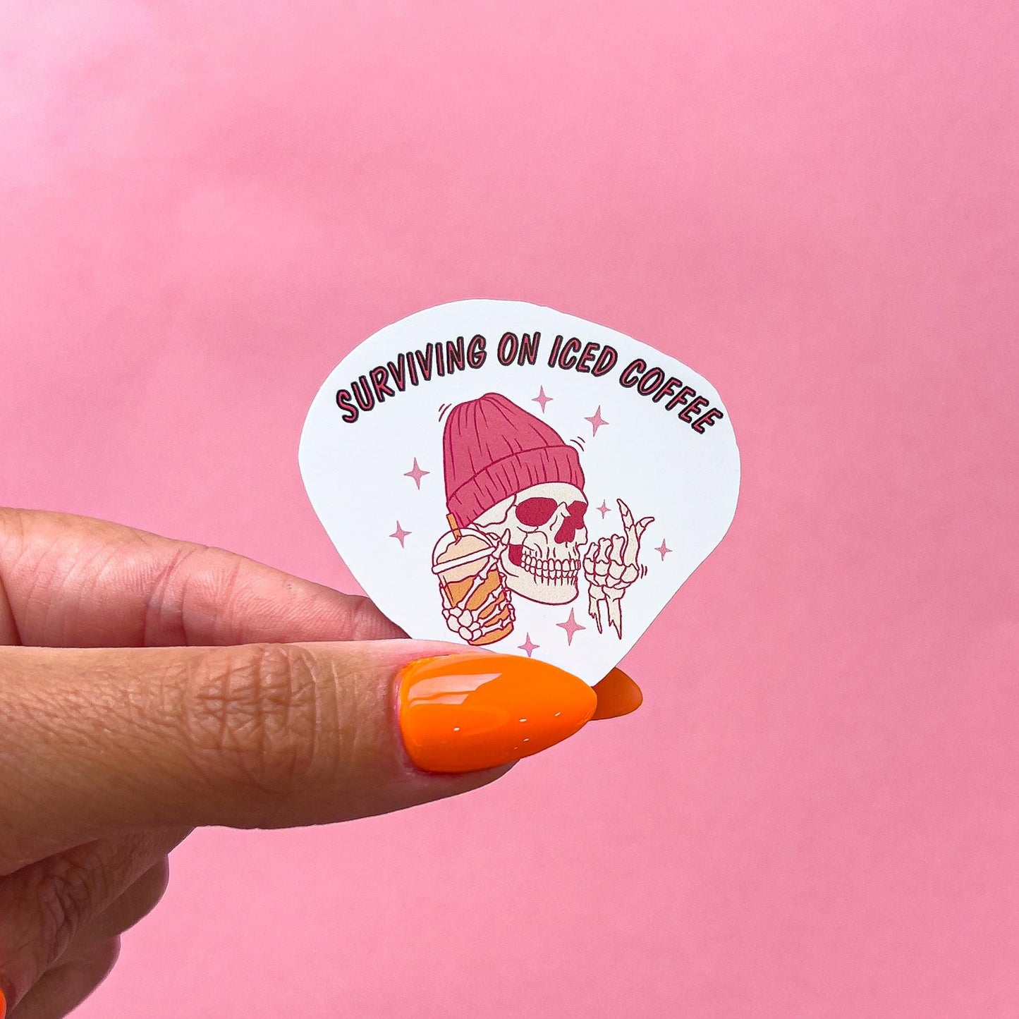 A vinyl decal sticker of a smiling skeleton wearing a pink colored beanie and holding a pink and orange iced coffee cup. The text reads "Surviving on iced coffee".