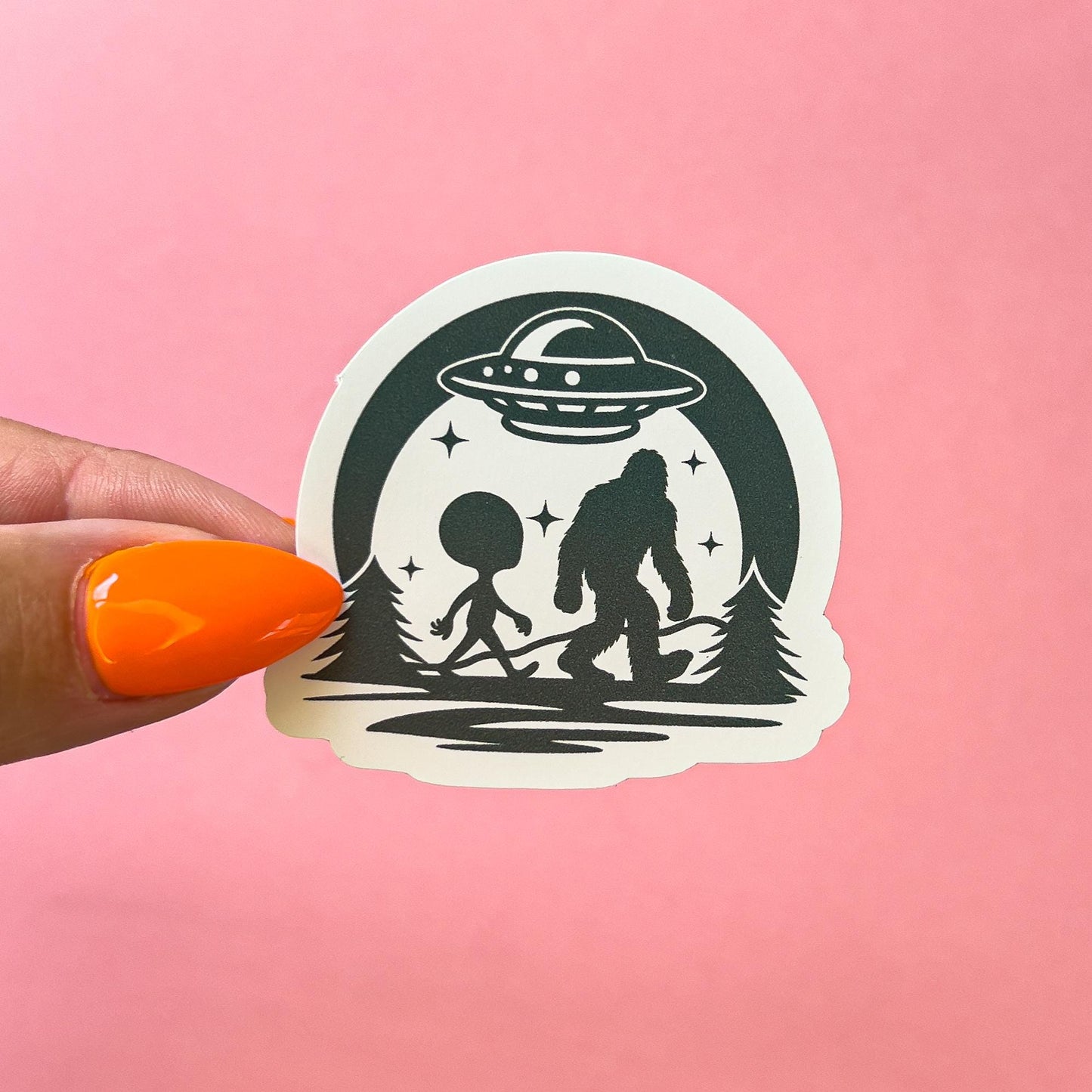 A vinyl decal sticker of a dark green silhouette of a Bigfoot and an alien walking side by side under a flying saucer. There is a moon and stars around the flying saucer.