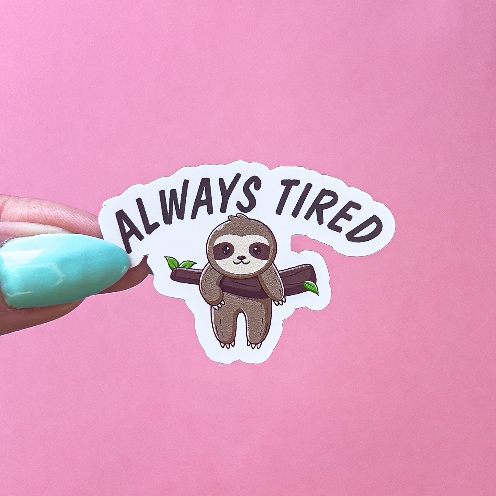 A vinyl sticker decal of a beige, brown, and white sloth laying over a tree limb with its legs dangling. Black text is curved at the top that reads “always tired”