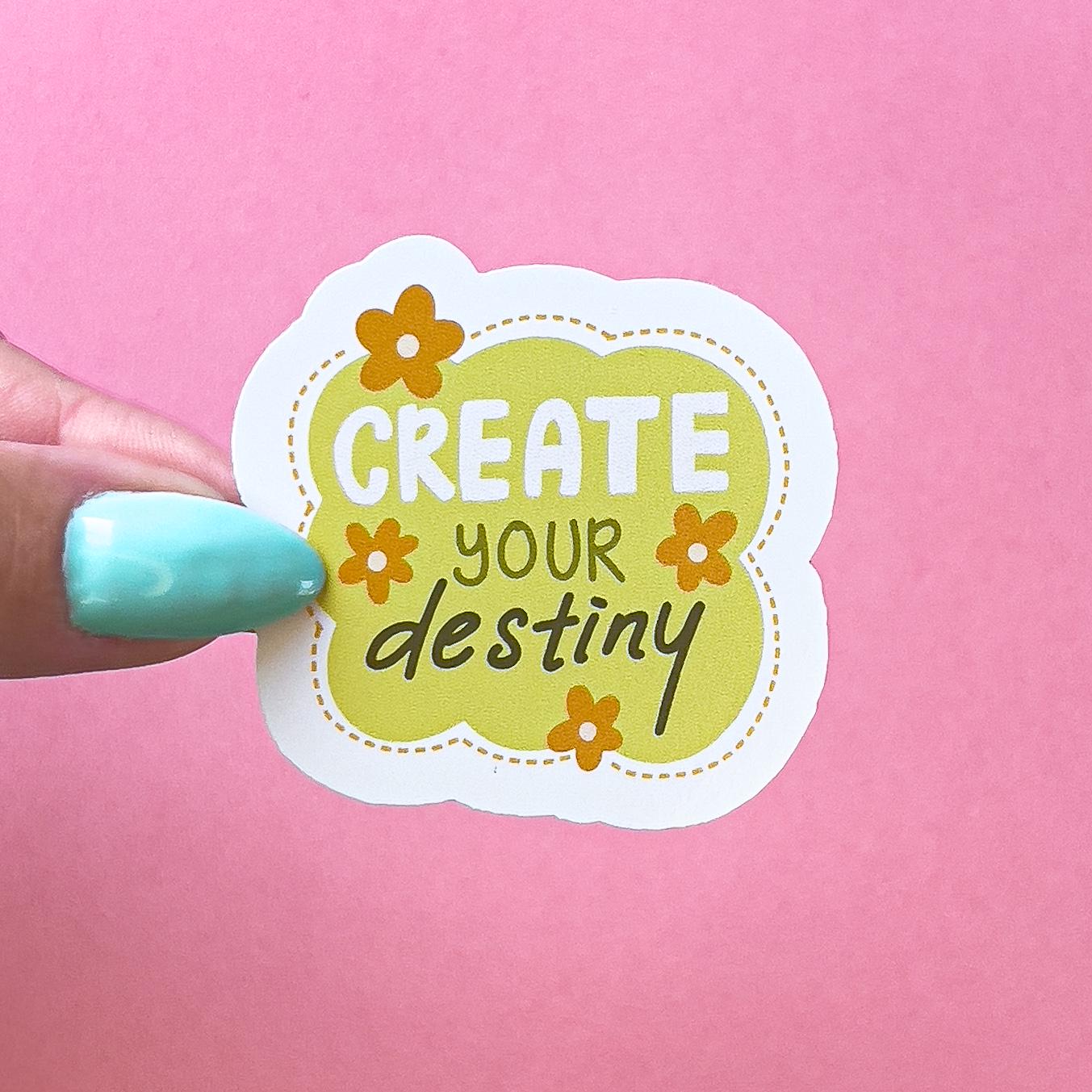 A vinyl decal sticker in an abstract shape with a yellow - green background encircled by a dotted line border. The text reads "Create your destiny" in white and green colors. There are four orange flowers around the text.