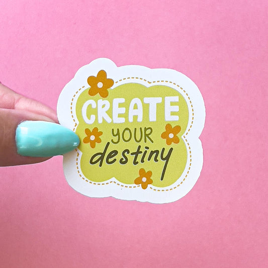 A vinyl decal sticker in an abstract shape with a yellow - green background encircled by a dotted line border. The text reads "Create your destiny" in white and green colors. There are four orange flowers around the text.
