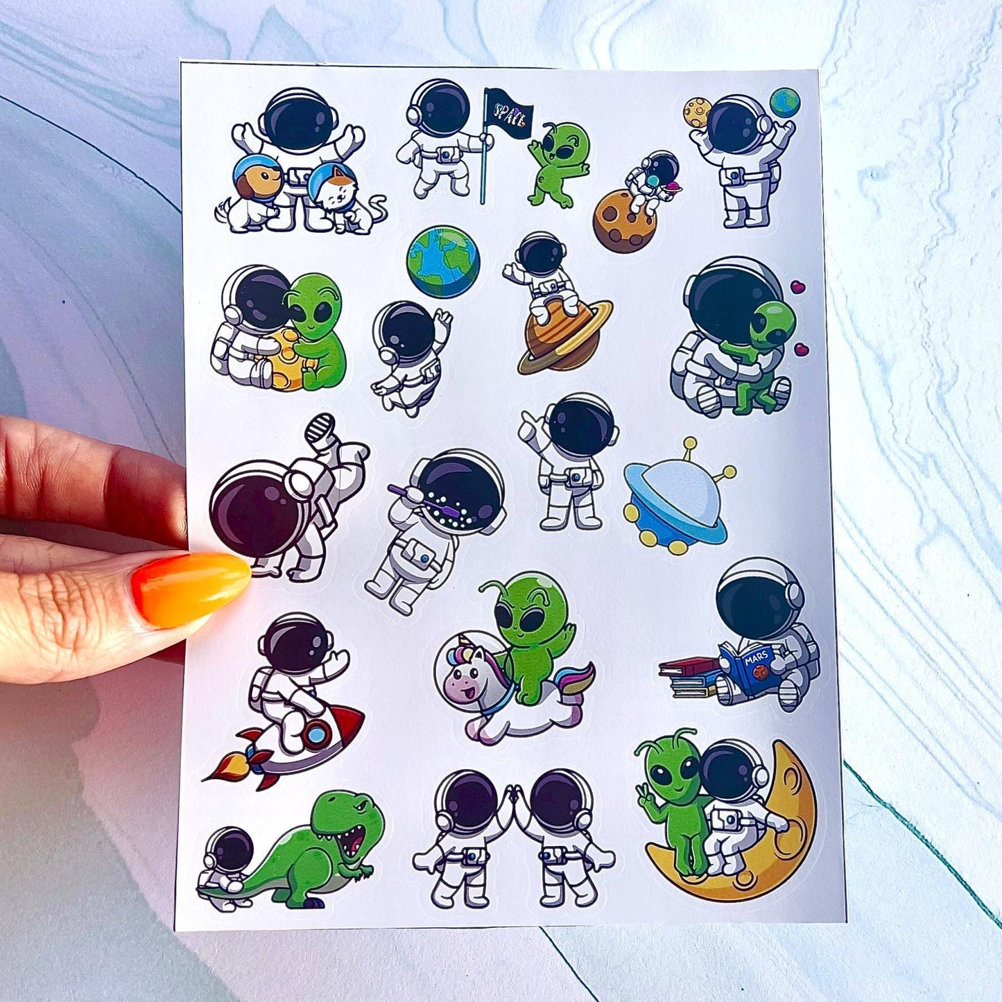 A sheet of colorful stickers featuring various space-themed characters, including astronauts, aliens, a dinosaur, a unicorn, a cat and dog, a Rocketship, a spaceship, and planets. The stickers are perfect for adding a fun and whimsical touch to any project.