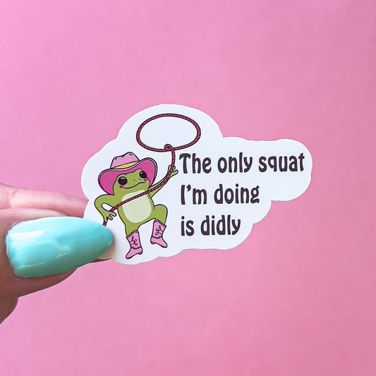 A vinyl decal sticker of a green frog wearing a pink cowboy hat and boots. The frog is holding onto a lasso. The text on the sticker reads 'The only squat I'm doing is diddly.'