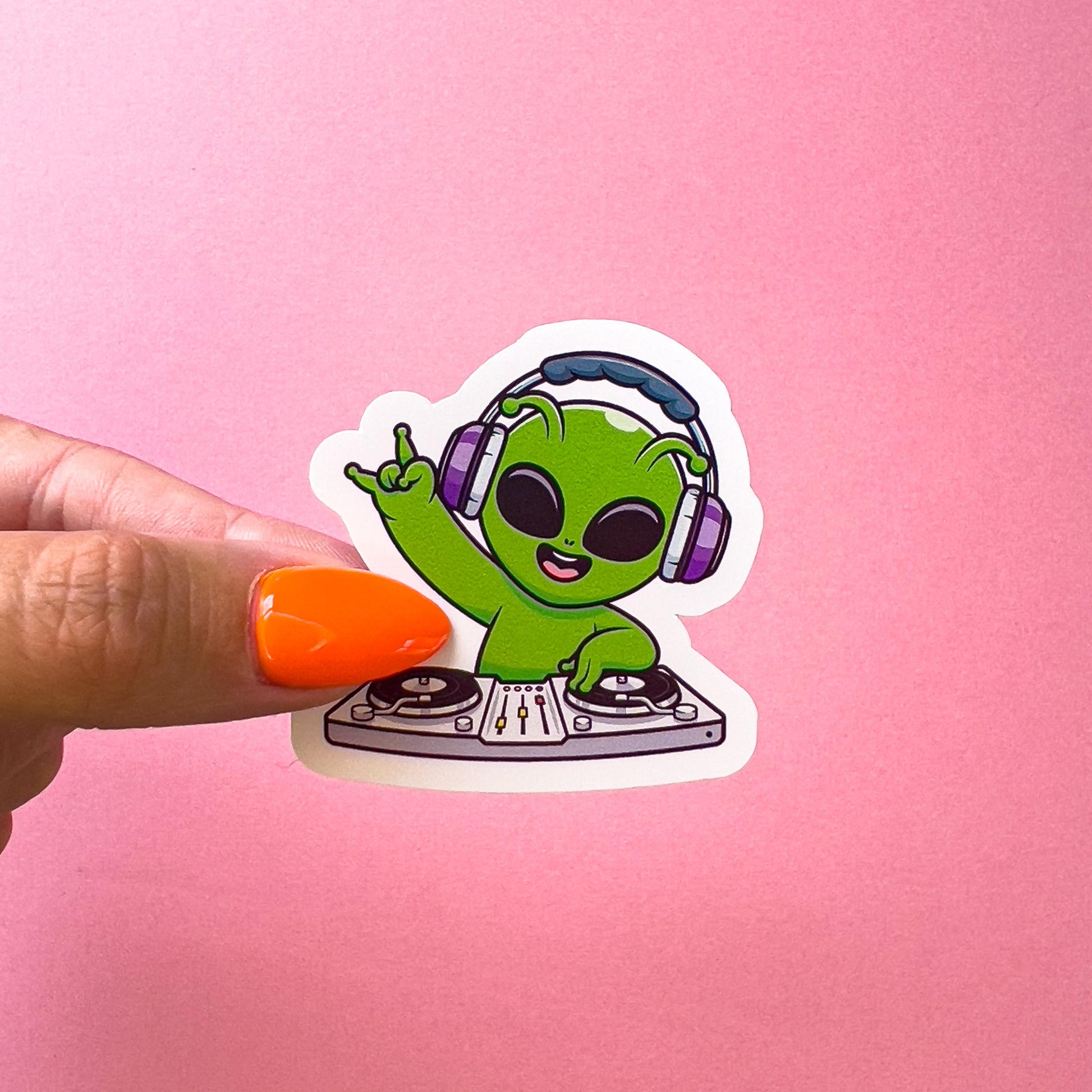 A vinyl decal sticker of a green alien wearing purple headphones and a toothy grin is playing a DJ mixer. The alien is holding its right hand up in the air with the index and pinky fingers extended. The mixer is gray with black knobs and a black record on each side.