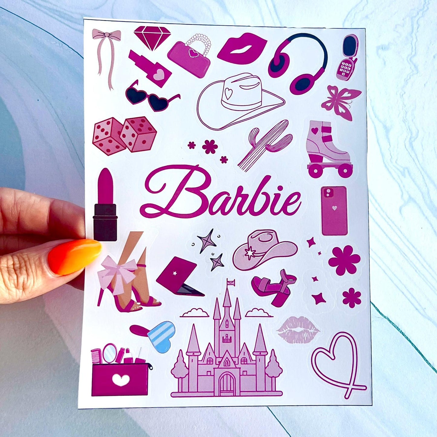 A sheet of pink and white stickers with a variety of images, including a cowboy hat, a castle, a pair of roller skates, a laptop, a pair of high heels, a lipstick, a pair of sunglasses, a heart, a cactus, a butterfly, a diamond, a purse, a pair of headphones, a cell phone, and dice. The word 'Barbie' is written in pink cursive letters at the top of the sheet.