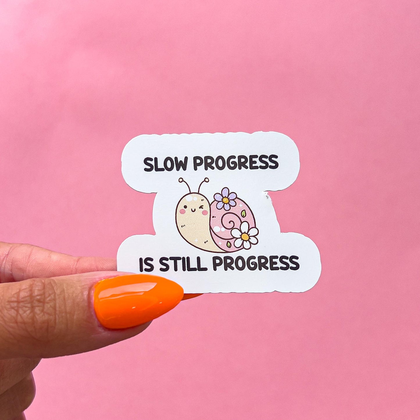 A vinyl decal sticker of a pink snail with flowers on its shell and pink blushed cheeks. The text on the sticker reads 'Slow progress is still progress.'