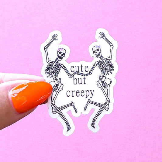 A vinyl decal sticker of two skeletons dancing and holding hands with the text 'cute but creepy' in between them. The skeletons are white and have black outlines. The text is in black. The skeletons are wearing pink and purple bows on their heads.