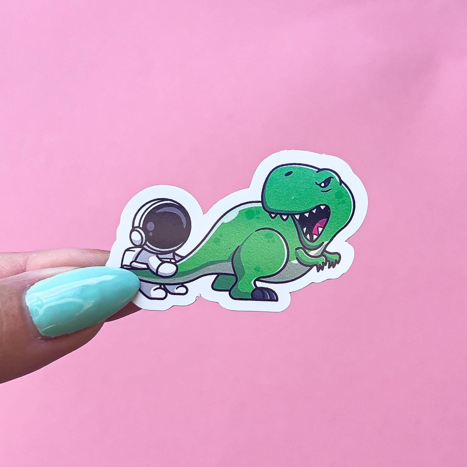 A vinyl decal sticker of an astronaut grabbing a dinosaurs tail. The dinosaur is a green, cartoon style T-Rex and it is looking back at the astronaut.