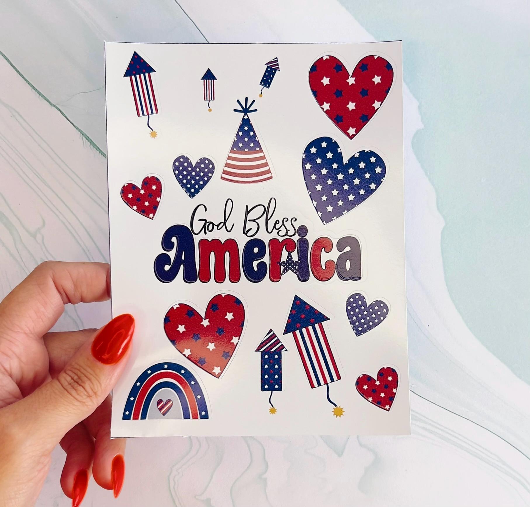 A sheet of various Fourth of July stickers. The stickers are of various shapes, including hearts, stars, fireworks, and a party hat. The text 'God Bless America' is written in a stylized font.