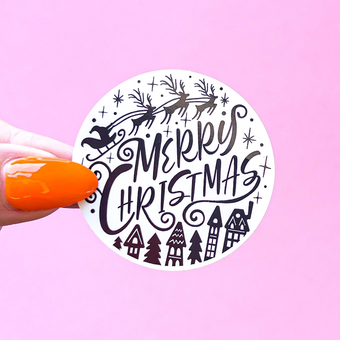 A white circle sticker with black 'Merry Christmas' text and a silhouette of Santa Claus in a sleigh pulled by reindeer. The sticker also has a small town scene with houses and trees.