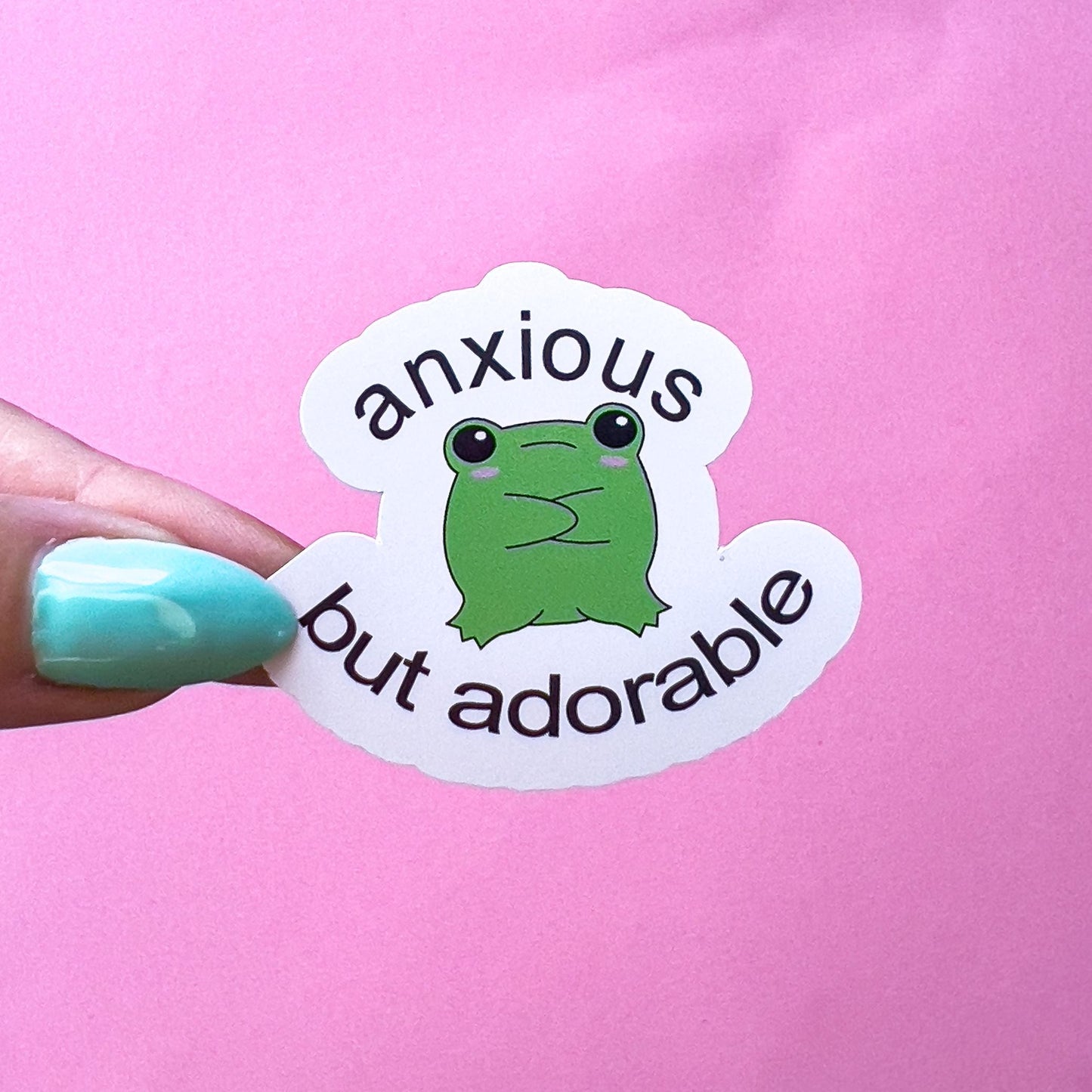 A vinyl decal sticker of a green cartoon kawaii frog with the text 'anxious but adorable'. The frog has pink blushed cheeks and is sitting with its arms crossed and its legs stretched out in front.