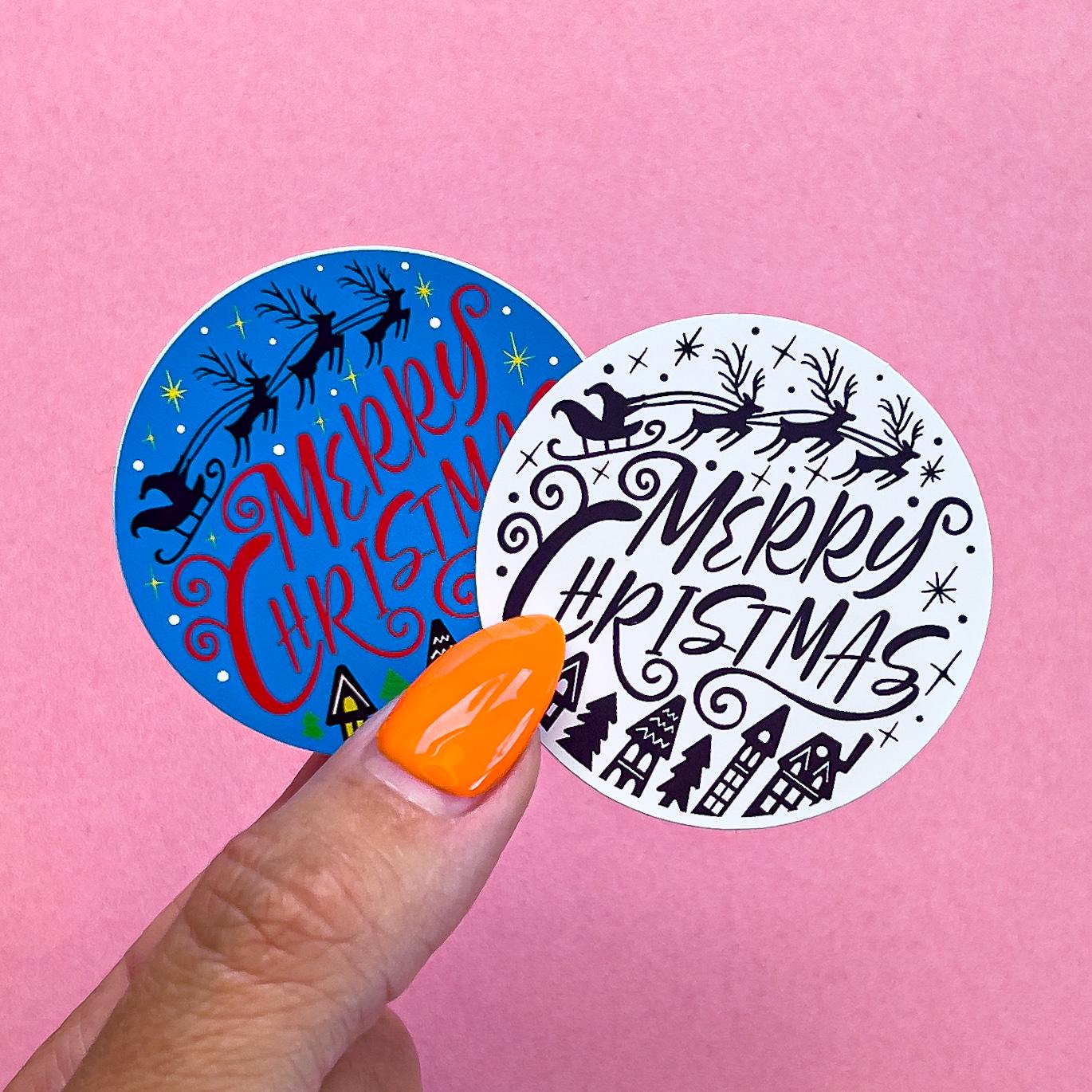 Two Christmas vinyl decal sticker options. One has a blue sky with “Merry Christmas” written accross in red letters. above the letters Santa’s sleigh is flying. along the bottom are homes lit up with yellow windows. The other Sticker is the same design, but black and white.