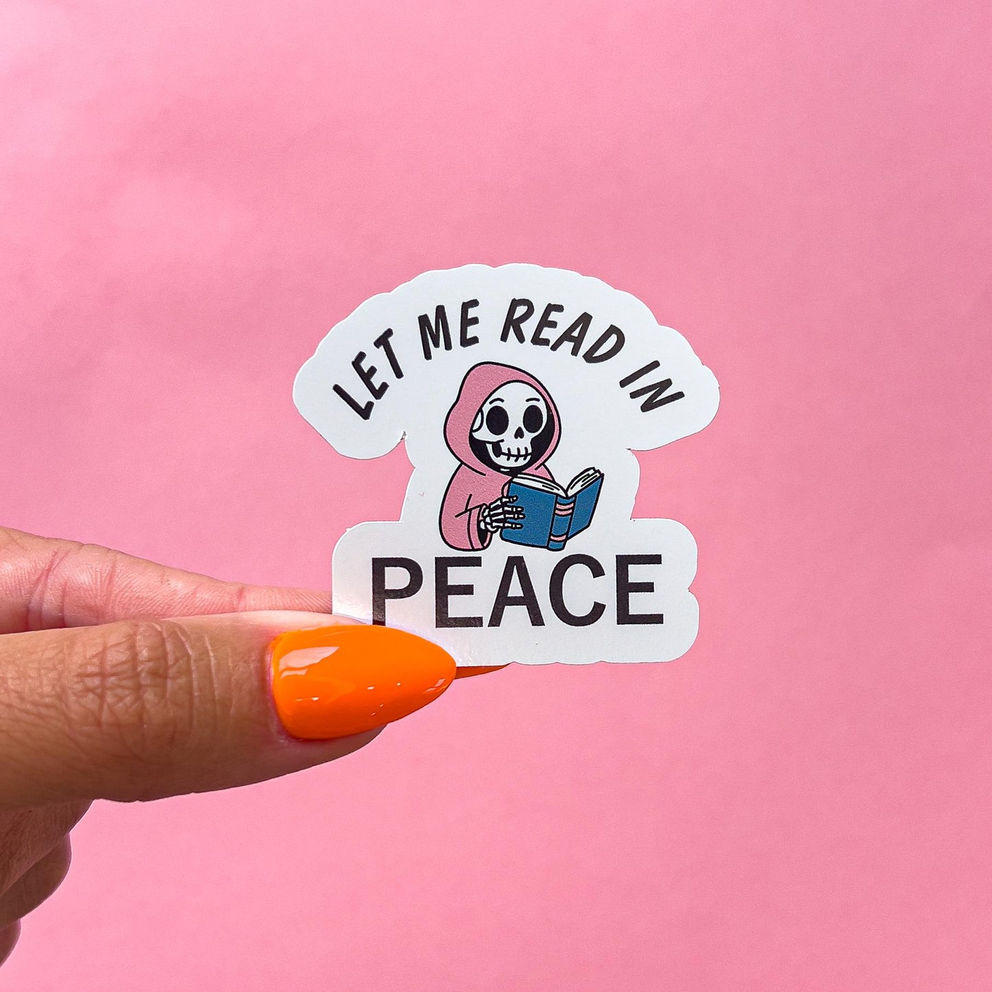 A vinyl decal sticker of a cartoon skeleton wearing a pink hooded cloak and reading a book. The text on the sticker reads 'Let me read in peace'.