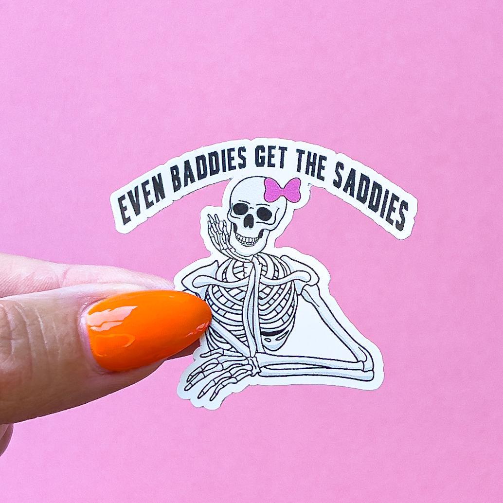 A vinyl decal sticker of a white skeleton with a pink bow on its head is sitting with its chin/ cheek resting on its hand. The text above the skeleton reads 'Even baddies get the saddies.'