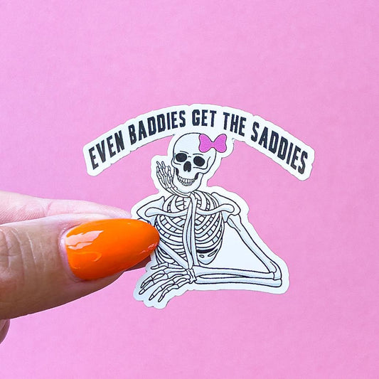 A vinyl decal sticker of a white skeleton with a pink bow on its head is sitting with its chin/ cheek resting on its hand. The text above the skeleton reads 'Even baddies get the saddies.'