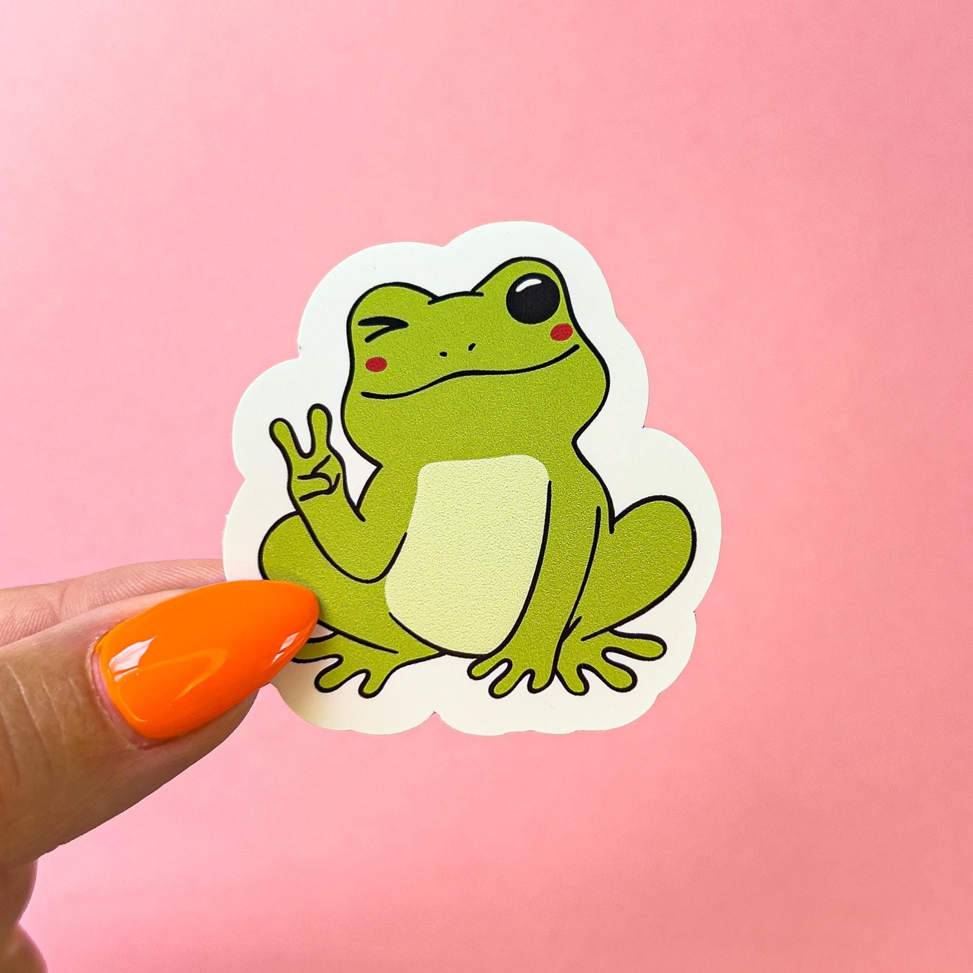 A vinyl decal sticker of a green, cartoon frog with blushed cheeks and holding up a peace sign.