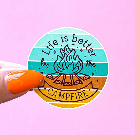 A round vinyl decal sticker with a blue, green, and orange striped background. The sticker has a campfire with flames in the center and the text 'Life is better by the campfire'.