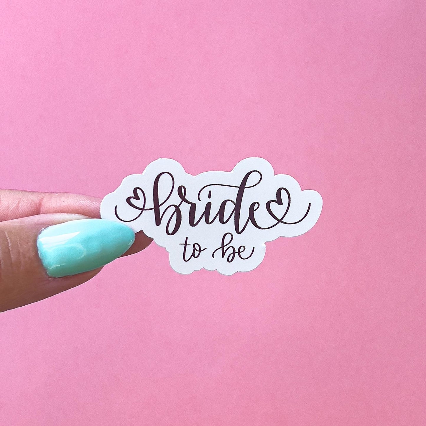 A white sticker with black cursive text that reads 'bride to be' with a heart on either side of the word 'bride'.