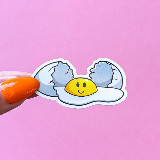 A vinyl decal sticker of a white cartoon egg with a cracked shell and a yellow smiley face yolk. The egg is in the shape of a fried egg with a white outline.