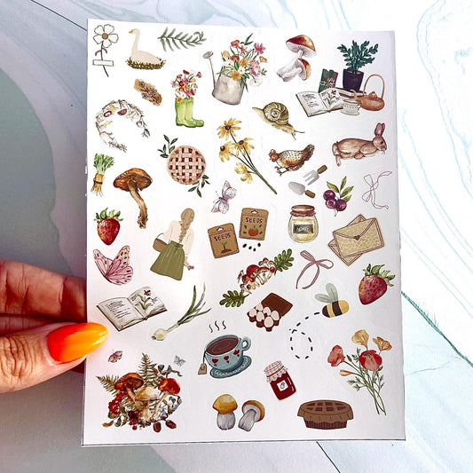 A sheet of cottagecore themed stickers. The stickers include pies, a jar of jam, mushrooms, vegetables, butterflies, a strawberry, tea, envelopes, recipe books, a chicken, a goose, flowers, plants, a bee, a rabbit, packs of seeds, a bow, a snail, ect