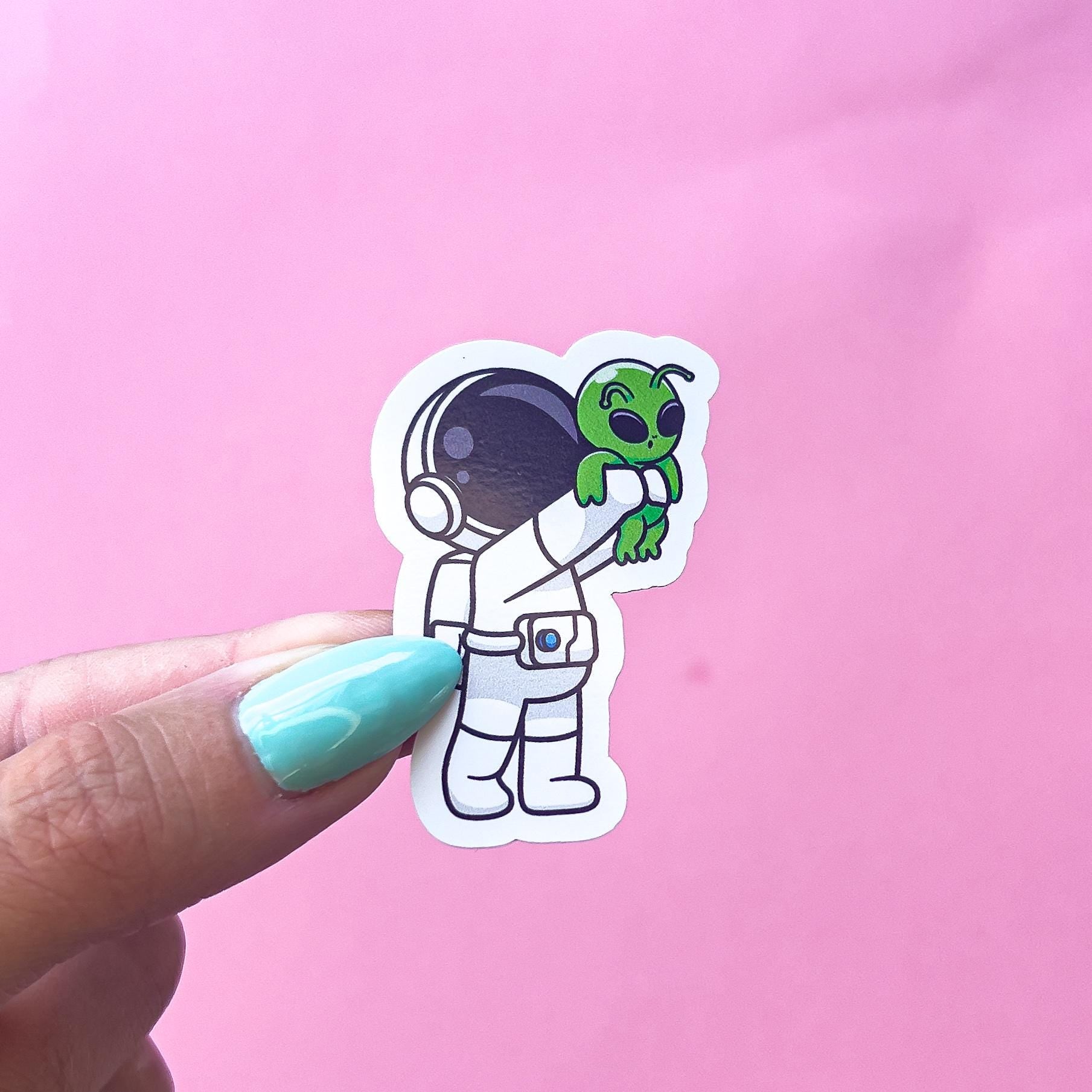 A vinyl decal sticker of an astronaut holding a green, baby alien in its arms. The astronaut is wearing a white spacesuit. The alien has large eyes and a small mouth.