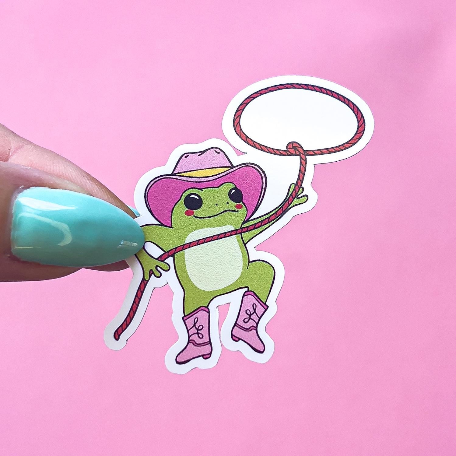 A vinyl decal sticker of a green frog wearing a pink cowboy hat and pink boots, holding a red lasso with a white circle at the end. The frog is swinging the lasso in the air.
