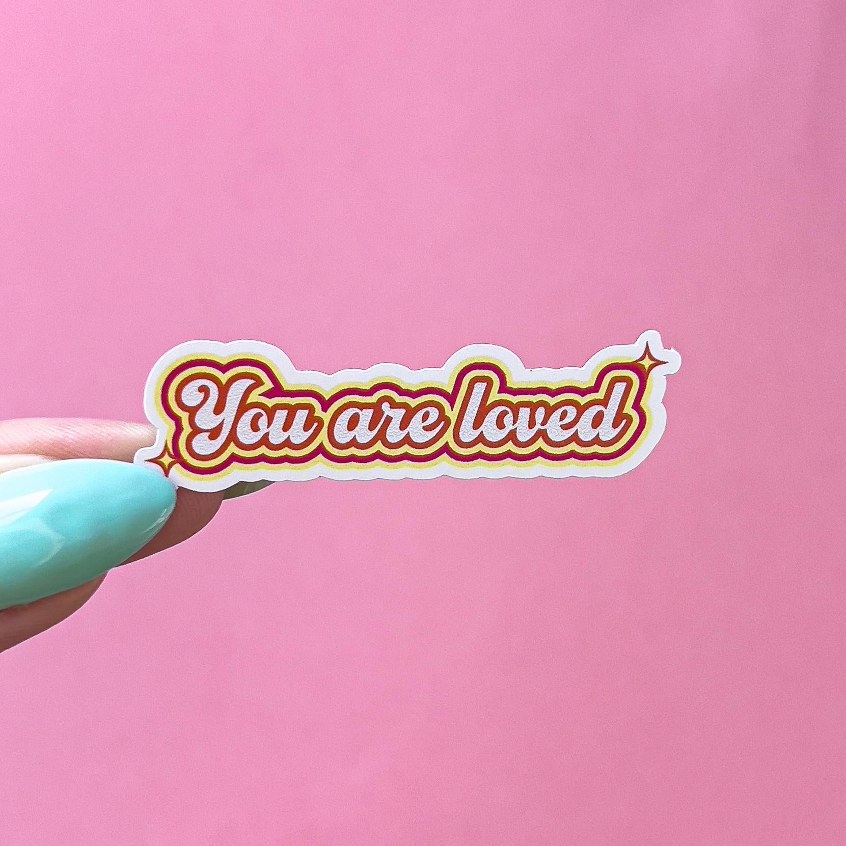 A vinyl decal sticker with a red and yellow outline. The sticker says 'You are loved' in a retro font style.