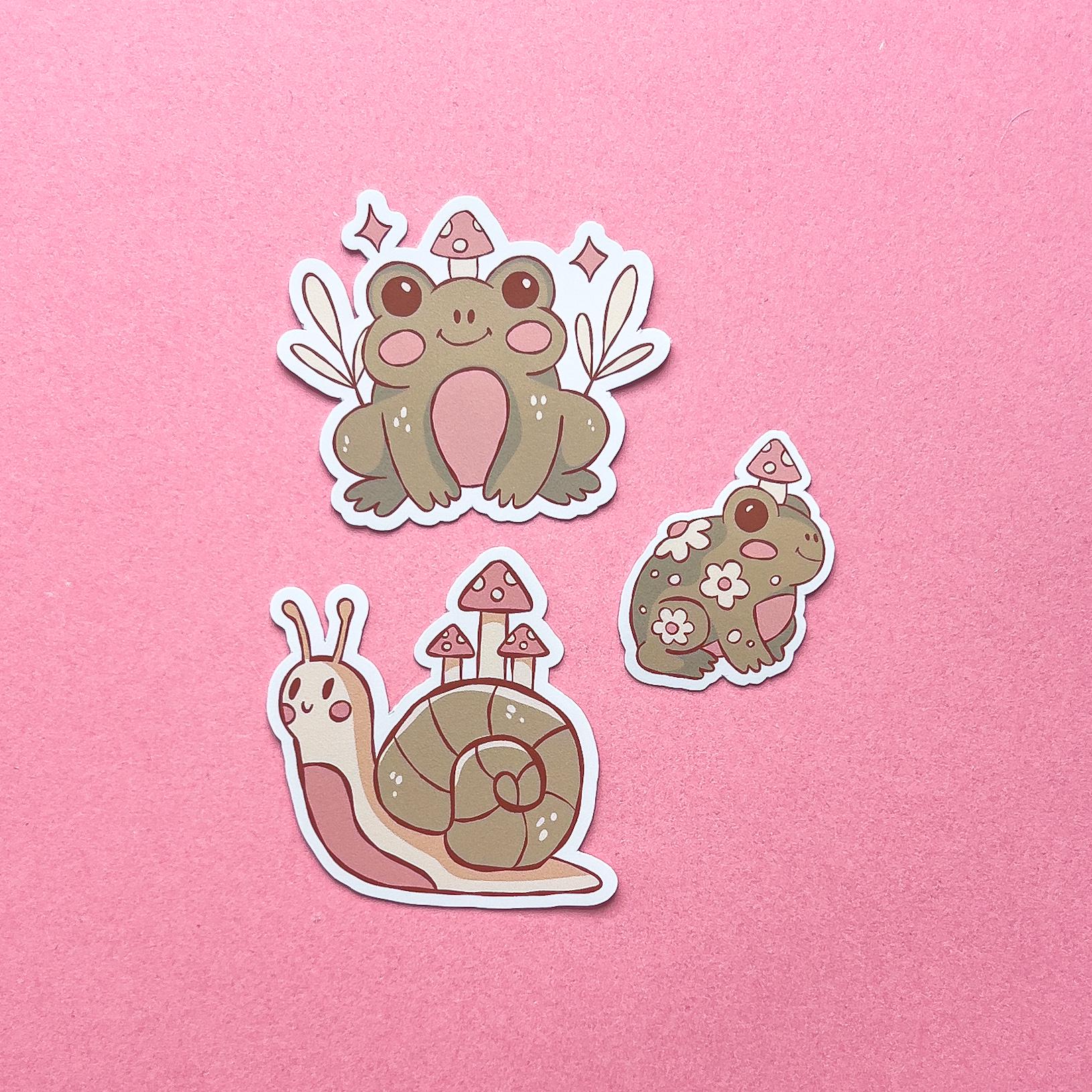 Three vinyl decal stickers of two cartoon frogs and a cartoon snail. Each have pink blushed cheeks, a small smile, and has mushrooms. All three are green, beige, and pink.
