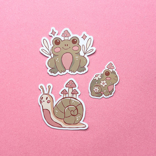 Three vinyl decal stickers of two cartoon frogs and a cartoon snail. Each have pink blushed cheeks, a small smile, and has mushrooms. All three are green, beige, and pink.