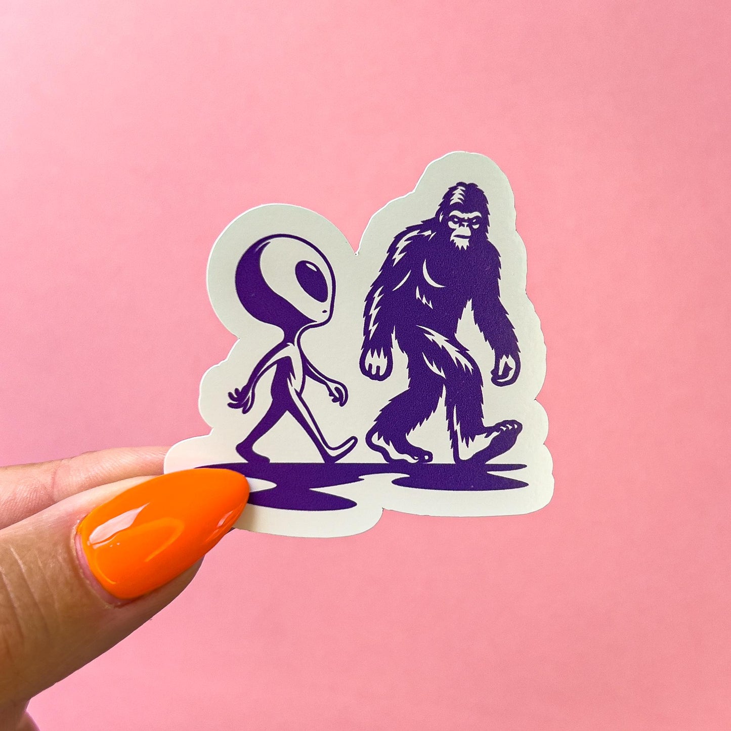 A vinyl decal sticker of an alien and big foot walking next to each other and looking at each other. Both the alien and big foot are in their typical form. They are dark purple silhouettes with minimal detail.