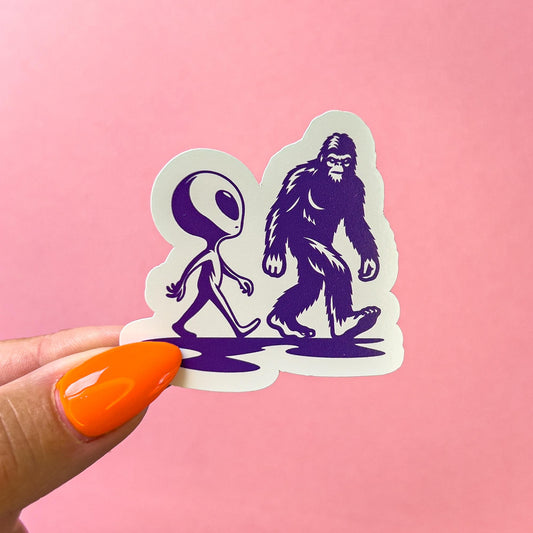 A vinyl decal sticker of an alien and big foot walking next to each other and looking at each other. Both the alien and big foot are in their typical form. They are dark purple silhouettes with minimal detail.