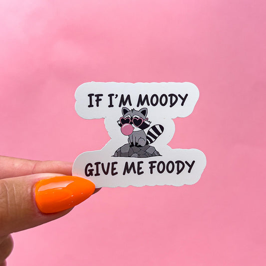 A vinyl decal sticker of a cartoon raccoon with pink sunglasses and blowing a pink bubblegum bubble. Black text reads 'If I'm Moody Give Me Foody.'