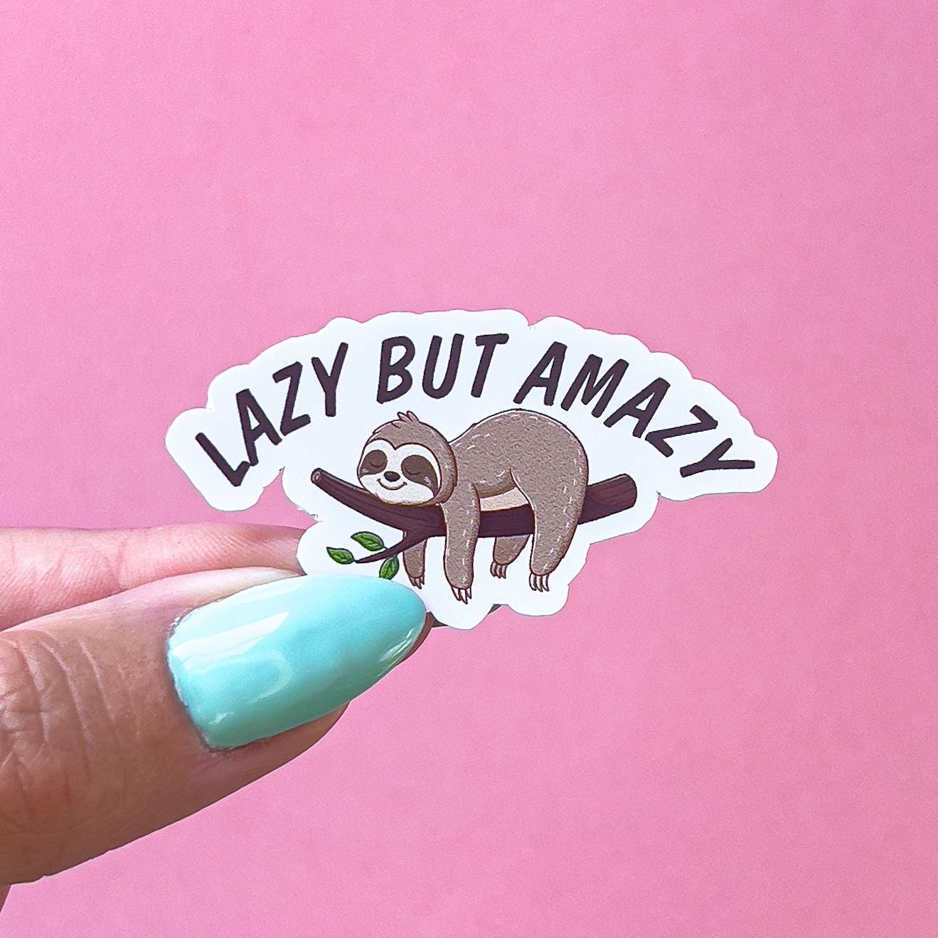 A vinyl decal sticker of a white, brown, and beige sloth laying on a tree limb with its limbs hanging under it. Underneath black text reads “lazy but amazy”.