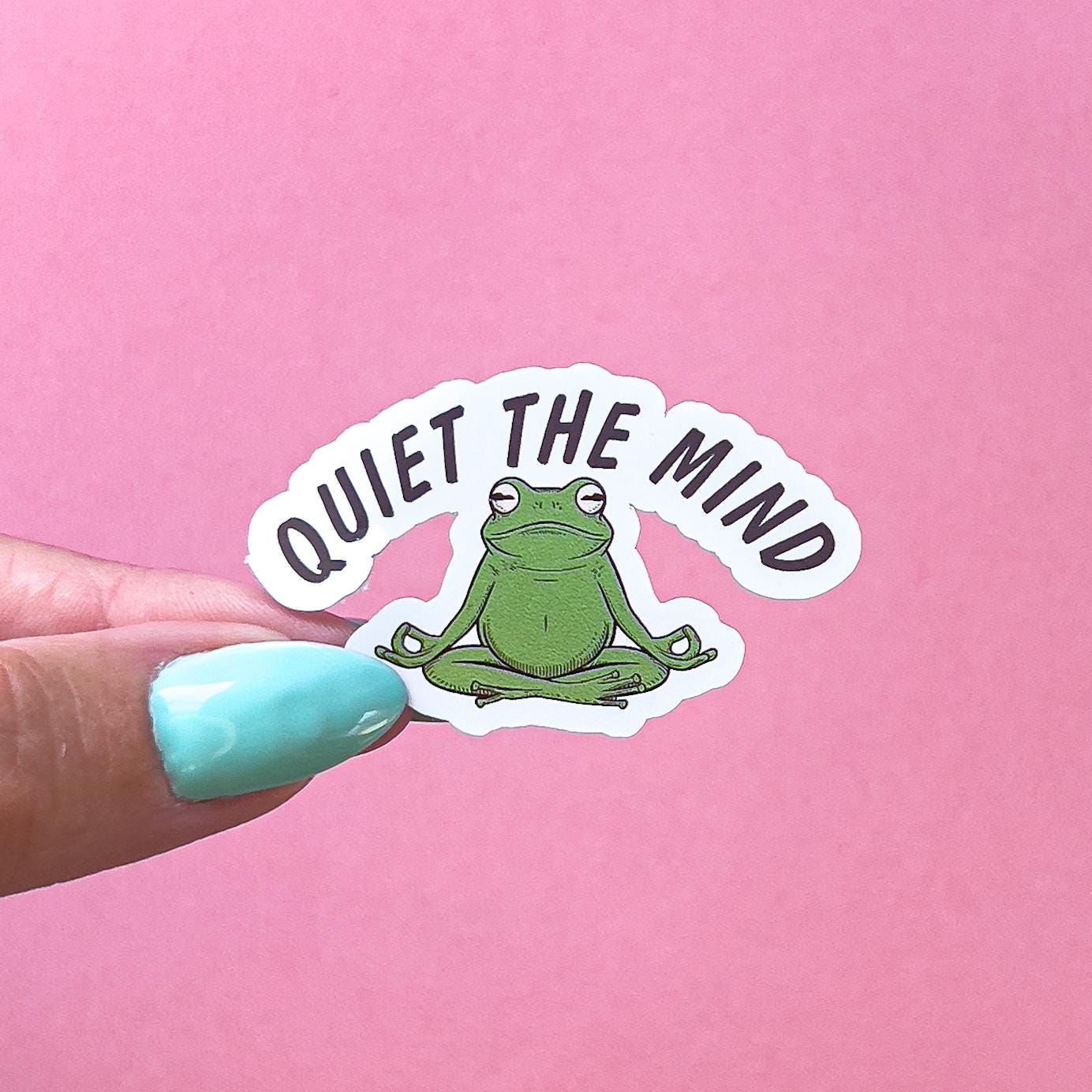 A vinyl decal sticker of a green frog  doing the Sukhasana, yoga pose. Above the frog black text reads “Quiet the Mind”