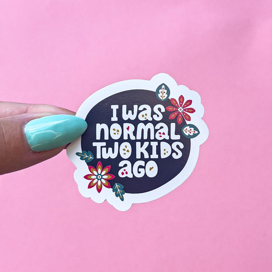 A vinyl decal sticker with a dark blue circle in the center. The circle has the text 'I was normal two kids ago' in white text. The circle is surrounded by colorful flowers.