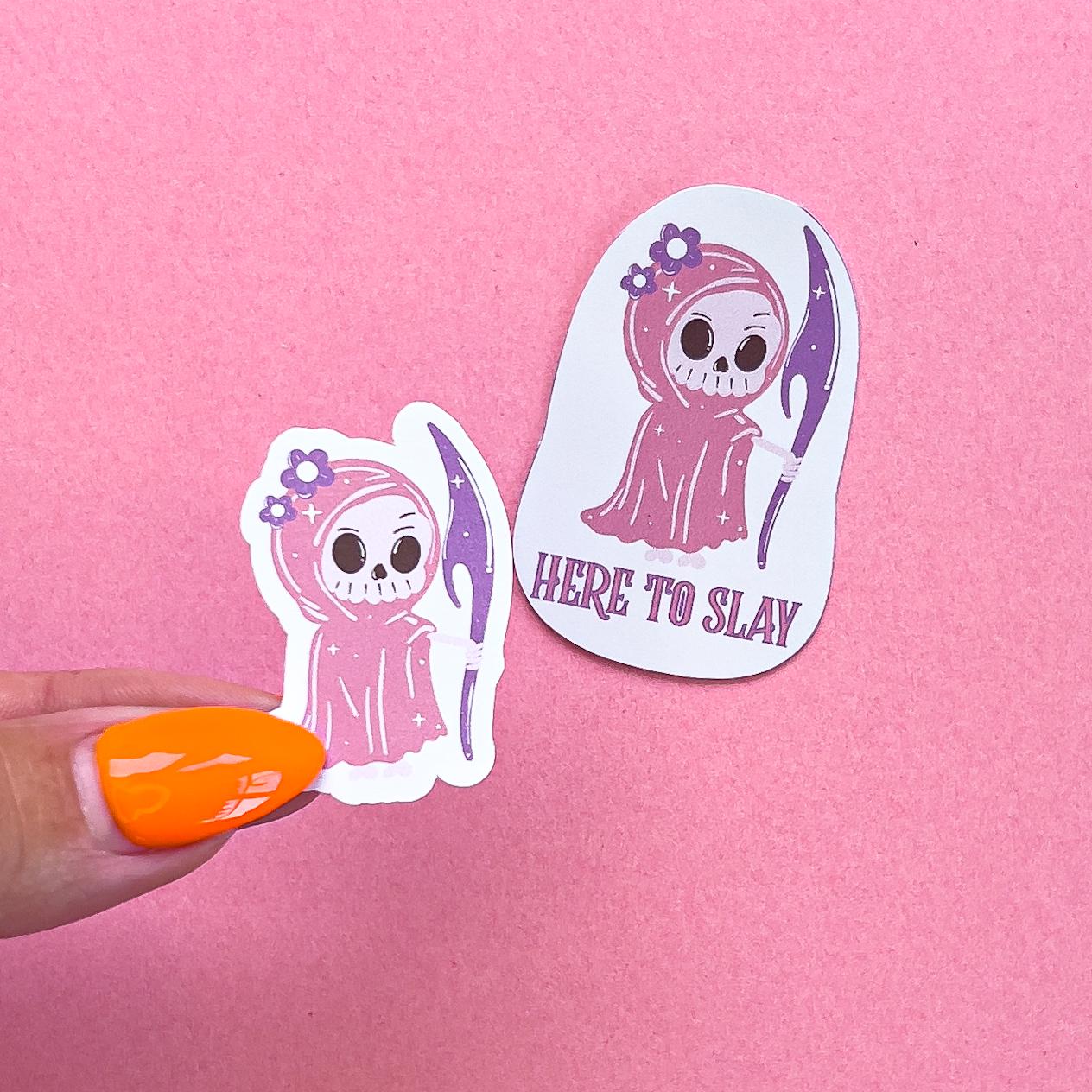 Two vinyl decal stickers of a girly grim reaper character. The grim reaper has a white skeleton face. She is wearing a pink cape/robe with purple flowers on the hood. She is holding a purple scythe. One sticker is just the girly grim reaper. The other sticker says "HERE TO SLAY" under the girly grim reaper.