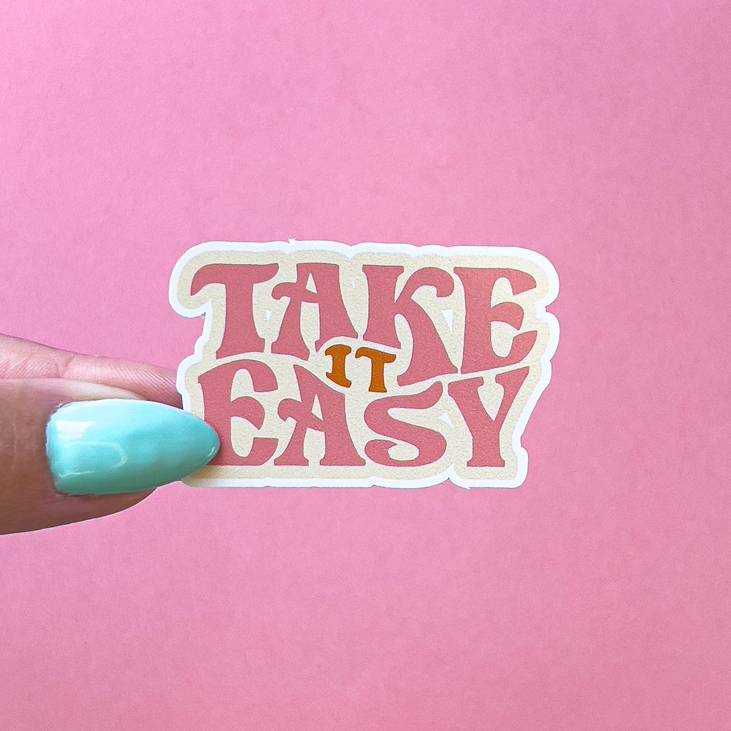 A vinyl decal sticker with the text 'Take it easy' in a retro font. The text is pink with a white outline and the word 'it' is in orange.